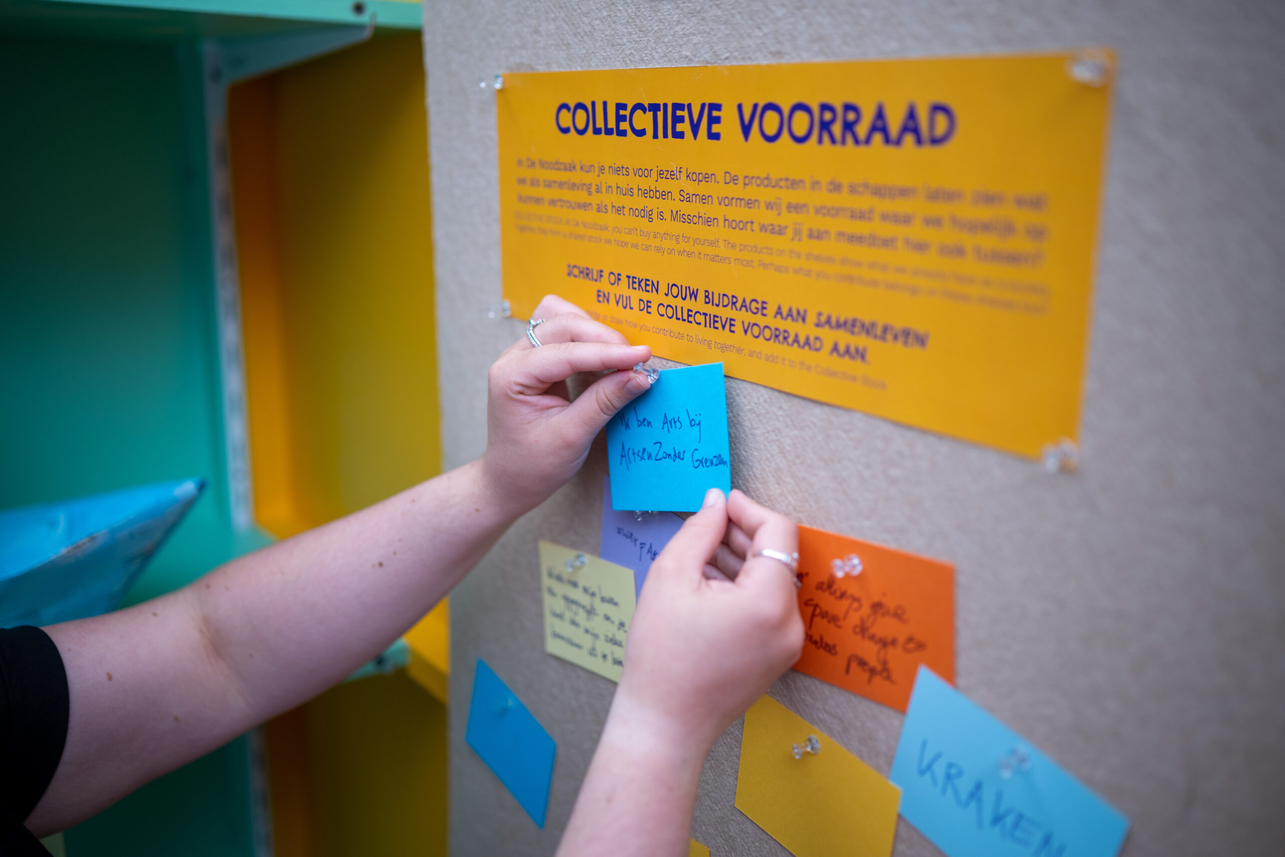 Handen hangen een briefje op een prikbord genaamd 'collectieve voorraad'. Op het briefje staat geschreven: "ik ben arts bij Artsen zonder Grenzen"