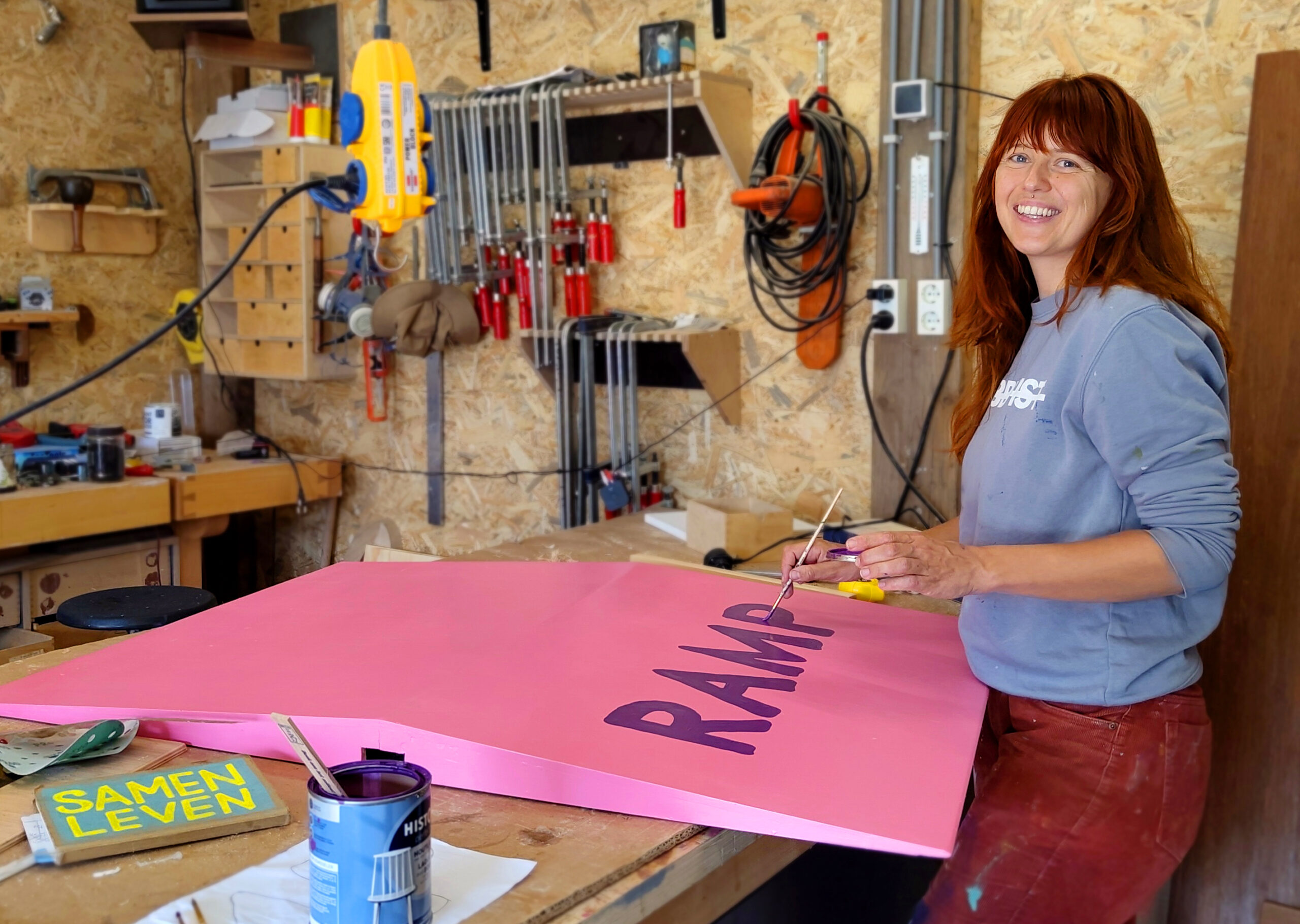 Maartje schildert met de hand de letters 'RAMP' op een roze rolstoeltoegankelijke oprit (ramp) in een houtwerkplaats.