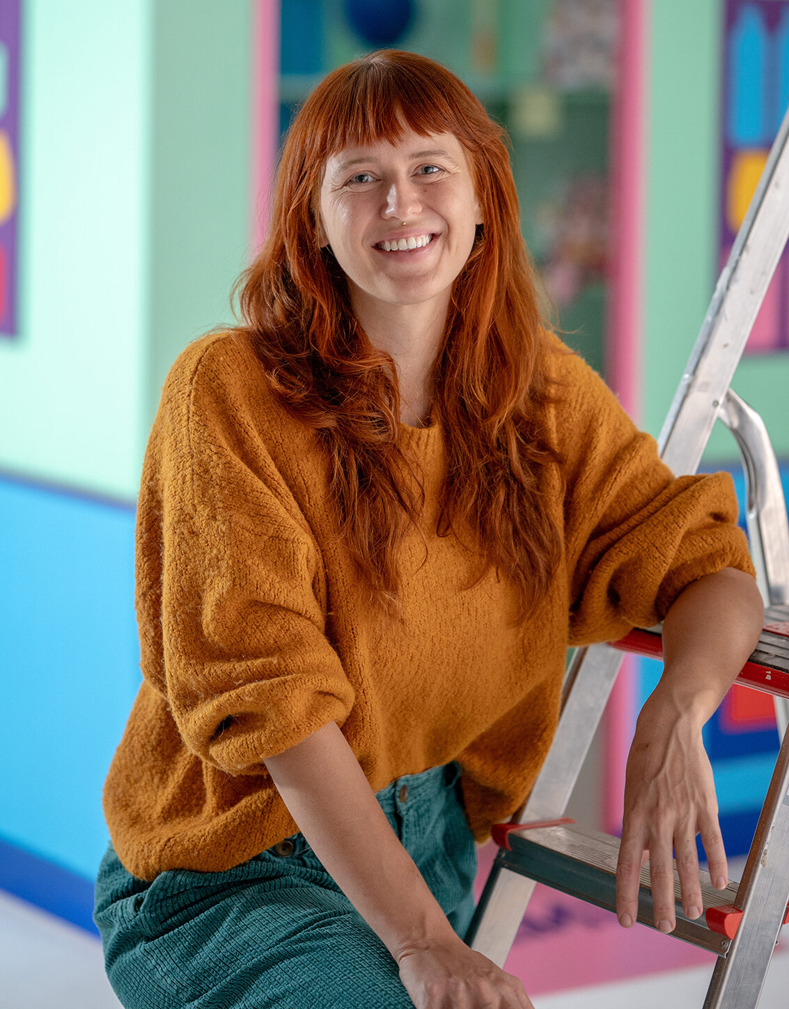 Portret van Maartje de Goede. Maartje poseert lachend tegen een ladder aan met als achtergrond De Noodzaak. Rood haar, gele trui, blauwe broek.
